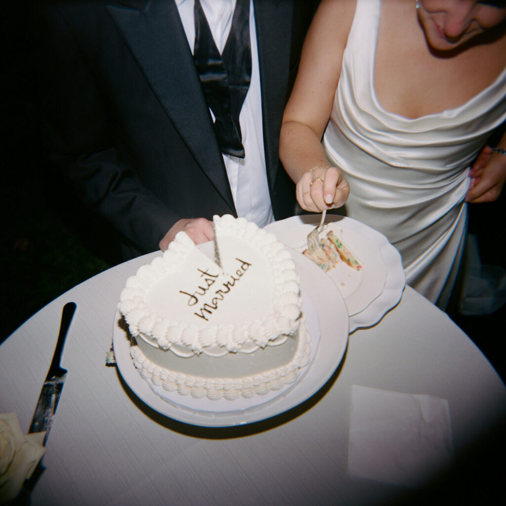 A bride and groom stand at a table cutting a heart-shaped cake that says "Just married." The bride is serving a slice onto a plate, showcasing the elegance of wedding catering Seattle couples adore.