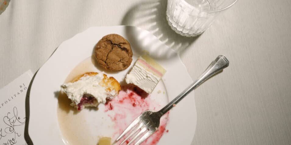 A white plate with dessert remnants, including cake, a cookie, and sauce, sits on a table next to a fork, a glass of water, part of a printed menu, and what appears to be a catering quote.