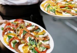 Two oval plates with colorful Caprese salads made of sliced tomatoes, mozzarella, basil leaves, and green olives, being held by a person wearing a white shirt and dark apron.