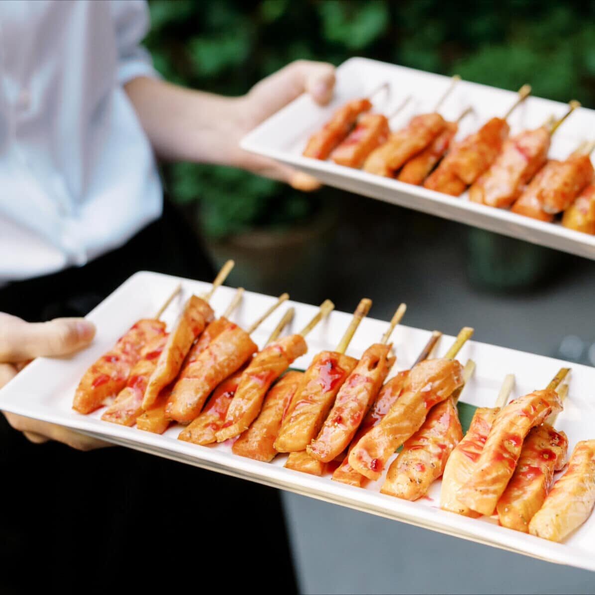 Two white rectangular plates are held by a person, each plate containing several skewers of grilled, glazed meat—perfect for drop off catering at your next event.