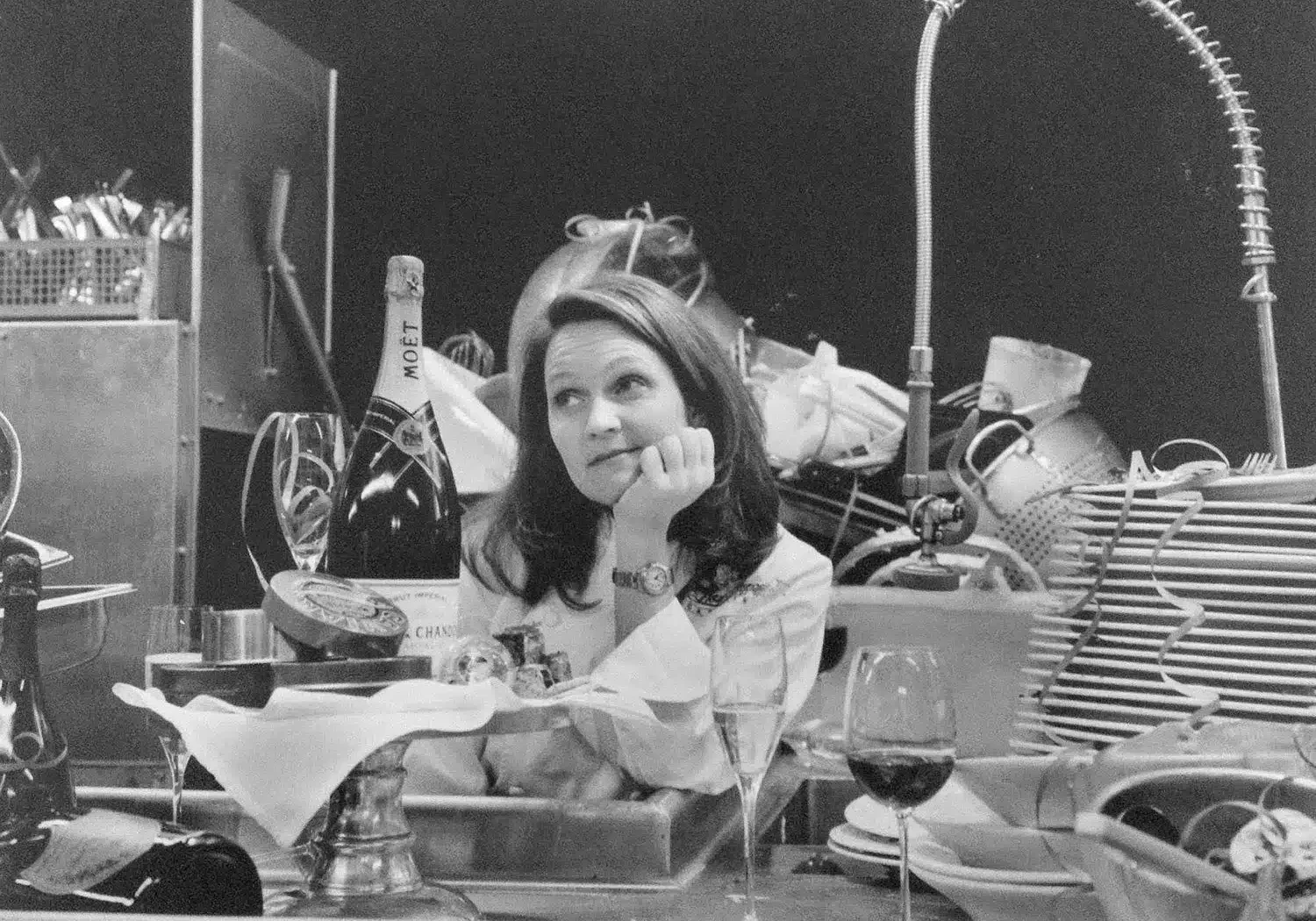 A woman sits at a cluttered kitchen counter, surrounded by dirty dishes, glassware, and a bottle of champagne—perhaps reflecting on a night catered by About Lisa Dupar Catering Seattle.