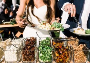 Two people in formal attire serve themselves food from a buffet with a variety of toppings and salads arranged in glass containers.