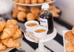 A small bottle of syrup and two white ramekins of jam are displayed on a pedestal, surrounded by plates of croissants.