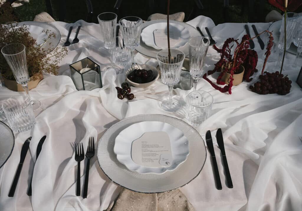 A table set with white plates, crystal glasses, black cutlery, and a white tablecloth, decorated with dried flowers, grapes, and a menu card on the central plate—perfect inspiration from top event vendors in Seattle.
