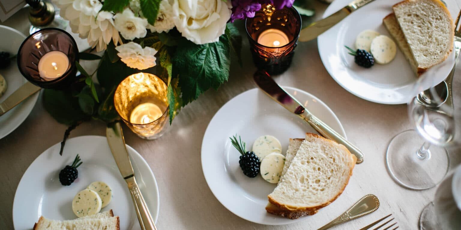 A table setting for wedding catering Seattle features plates with slices of bread, herb butter, and a blackberry garnish, surrounded by candles, silverware, wine glasses, and an elegant floral centerpiece.