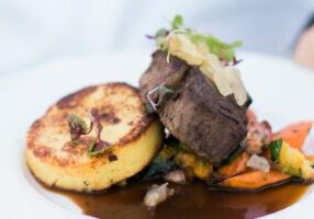 A plated meal featuring a seared round of polenta, a portion of cooked beef topped with garnish, vegetables, and a brown sauce.
