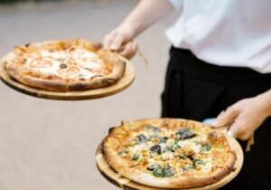 A person wearing a white shirt is holding two wooden trays with different pizzas, one topped with tomatoes and the other with greens and mushrooms.