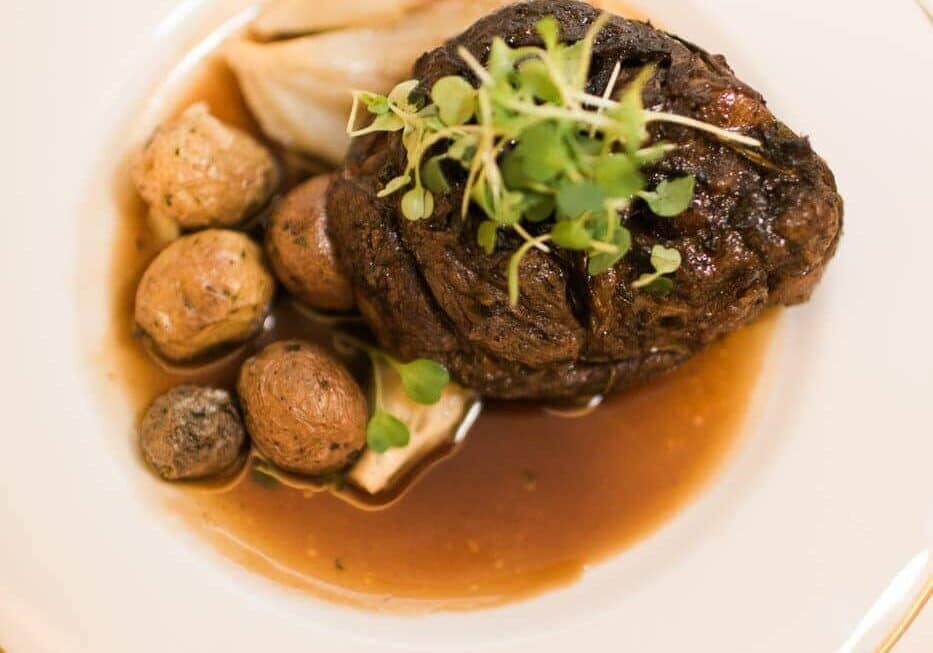 A plated dish featuring a cooked beef steak topped with microgreens, small roasted potatoes, and a brown sauce.