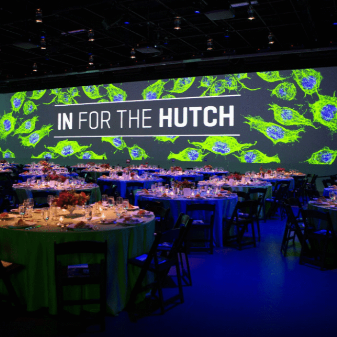 A banquet hall with round tables set for an event, featuring a large screen displaying "IN FOR THE HUTCH" over a background of green and blue cell images.