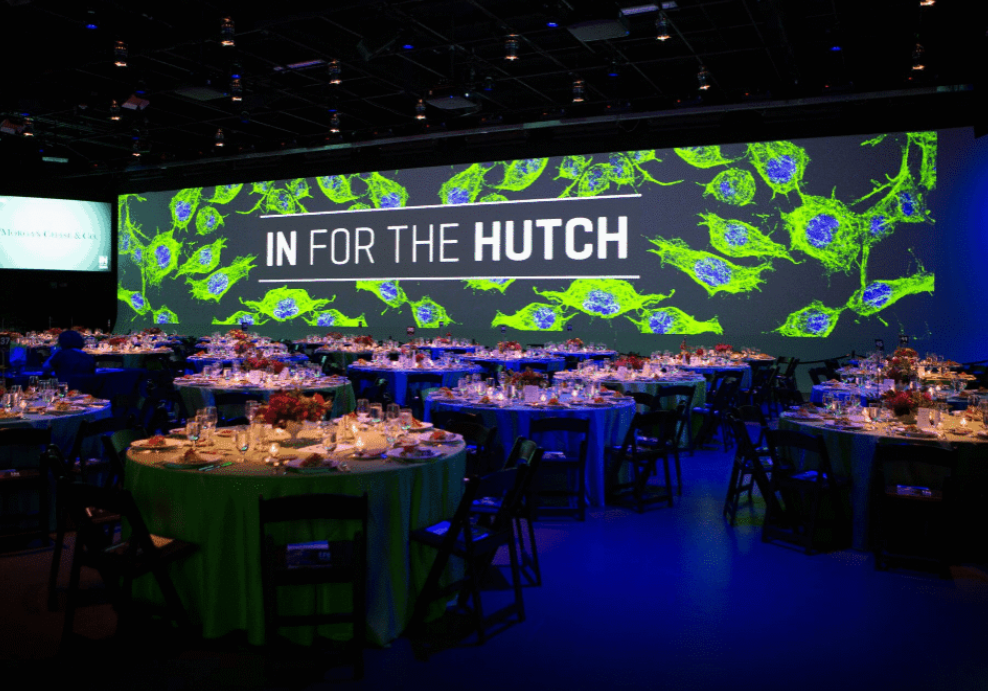 A banquet hall with round tables set for an event, featuring a large screen displaying "IN FOR THE HUTCH" over a background of green and blue cell images.