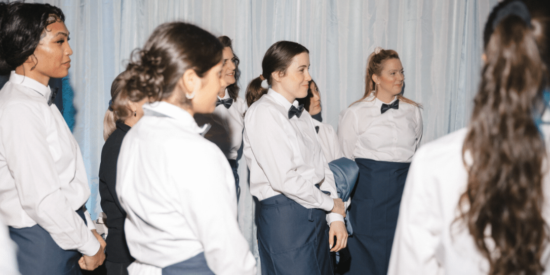 A group of people wearing white shirts, dark aprons, and bow ties stand together in front of a light-colored curtain, appearing to listen attentively—highlighting the professionalism found in careers at Lisa Dupar Catering.