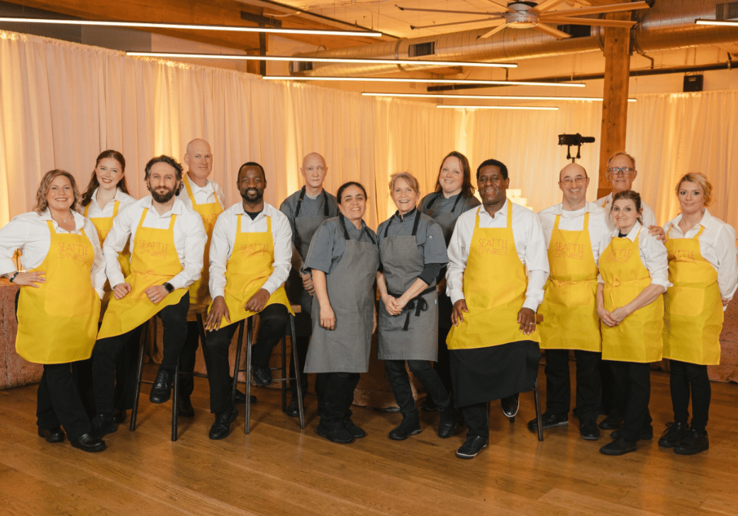 A group of thirteen adults in uniform aprons, standing and sitting indoors on a wooden floor, pose for a group photo in front of cream curtains—showcasing the team spirit behind rewarding careers at Lisa Dupar Catering.
