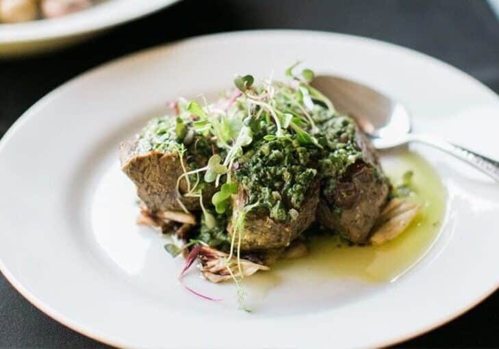 A white plate with two pieces of cooked meat topped with green herb sauce and microgreens, served with a spoon.