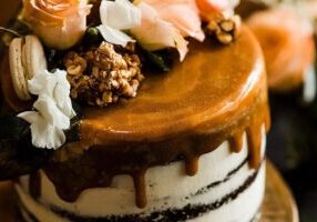 A layered cake with caramel drip topping, decorated with pink roses, white flowers, walnuts, and a macaron.