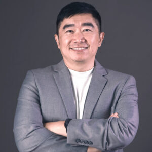 Man in a grey suit jacket and white shirt stands with arms crossed, smiling at the camera against a plain dark background.
