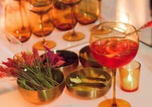 A table with a red cocktail in a wine glass, garnished with a straw, surrounded by metal bowls of garnishes, dried flowers, and amber glassware.
