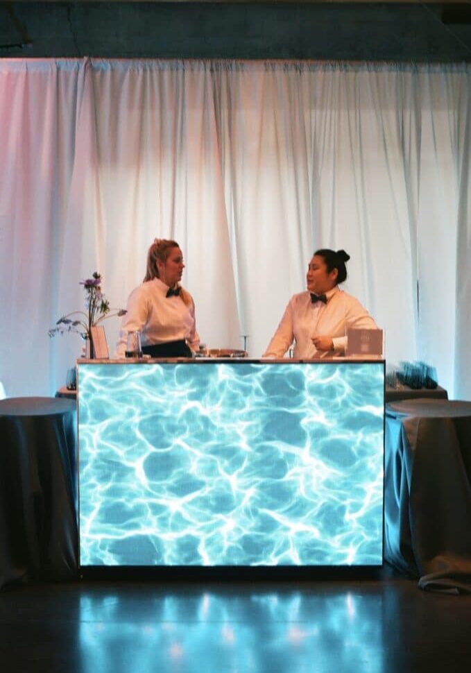 Two people in white shirts stand behind a bar counter with a blue water-like light display, set against a white curtain backdrop—an inviting scene that reflects the vibrant team spirit found in careers at Lisa Dupar Catering.
