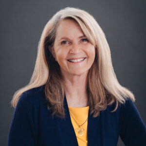Smiling woman with long blonde hair wearing a navy blue blazer over a yellow top, posing against a plain dark background.