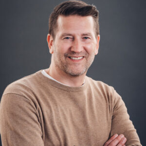 A man with short brown hair and light stubble, wearing a beige sweater over a white shirt, smiles with arms crossed against a plain dark gray background.