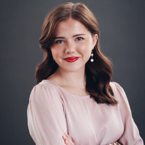 A woman with wavy brown hair, red lipstick, and pearl earrings, wearing a light pink blouse, stands with arms crossed against a dark gray background.