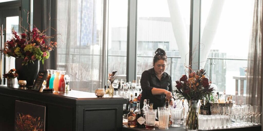 A bartender prepares drinks behind a black bar setup with bottles, glassware, and floral arrangements in a modern venue with large windows—discover more about joining this team through Careers at Lisa Dupar Catering.