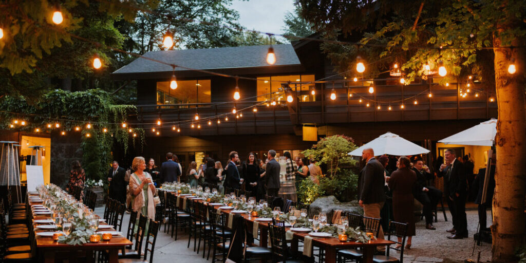 Outdoor evening event with guests mingling near decorated dining tables under string lights, surrounded by trees and patio umbrellas, enhanced by exceptional wedding and event catering.