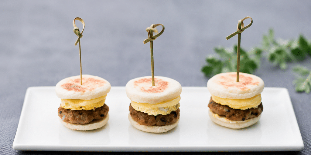 Three mini breakfast sandwiches with egg and sausage are arranged in a row on a rectangular white plate, each held together with a decorative toothpick—perfect for your next drop off catering event.